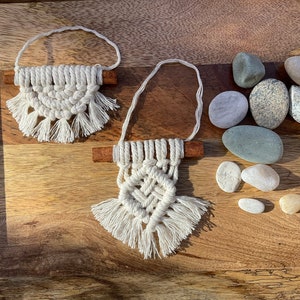 May include: Two small macrame wall hangings with wooden dowels and fringe, displayed on a wooden surface with smooth gray and white stones.