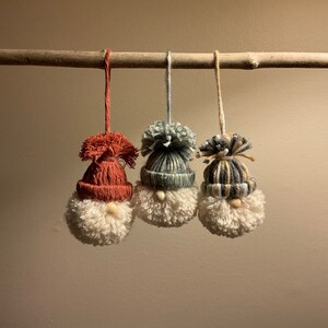 May include: Three handmade gnome ornaments hanging from a branch. The gnomes are made of yarn and have white pom-pom beards. Each gnome is wearing a different colored hat: red, blue and white striped, and yellow and white striped.
