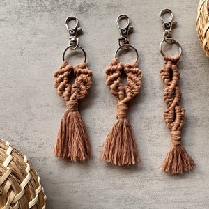 May include: Three brown macrame keychains with silver key rings and tassels. The keychains are made with a knotted rope design.