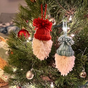 May include: Two handmade gnome ornaments hanging on a Christmas tree. One gnome has a red knit hat and the other has a blue knit hat. Both gnomes have white pom-pom beards.