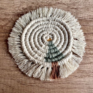 May include: A round, white macrame coaster with a green and brown tree design in the center. The coaster has a fringe border.
