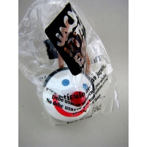 May include: A white, round toy with a red smile, blue eyes, and a black and white tag. The toy is inside a clear plastic bag. Text on the toy reads "artículo" and "No debe usarse".