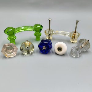May include: A collection of colorful cabinet hardware, including green, clear, blue, and white knobs and pulls. The hardware features various shapes, including faceted and round designs, with metal accents.