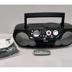 May include: A black Philips boombox with a CD player, speakers, and a handle. A remote control and instruction manual are also present. The boombox has a digital display and various control buttons. The speakers have a silver mesh covering.