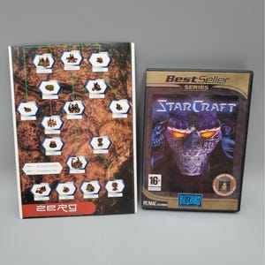 May include: A game box for StarCraft, a science fiction real-time strategy video game, with a dark cover featuring a menacing alien face. Next to it is a game guide with a brown and white map and the word "zerg".