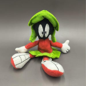 May include: A plush toy of Marvin the Martian, featuring a green helmet, red body, and white gloves and shoes. The toy has black eyes and a green skirt. The toy is a character from the Looney Tunes cartoon series.