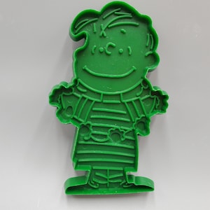 May include: Green plastic cookie cutter shaped like a cartoon character wearing a striped shirt. The character has a round head with a smile and a simple design.