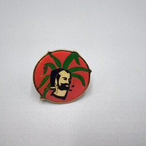 May include: A round, red and gold pin featuring a bearded man smoking a pipe, with green leaves in the background. The man's face is in profile, with black hair and a black beard. The pin has a gold border.