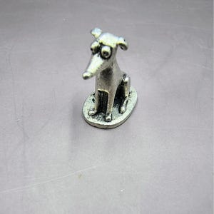 May include: A small, silver-colored dog figurine with oversized eyes and a long snout. The dog is sitting on an oval base. The figurine has a whimsical, cartoonish design.