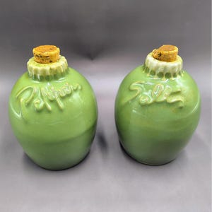 May include: Two green ceramic spice jars with cork stoppers. One jar is labeled "Pepper" and the other "Salt" in a raised, cursive font. The jars have a bulbous shape and a decorative rim.