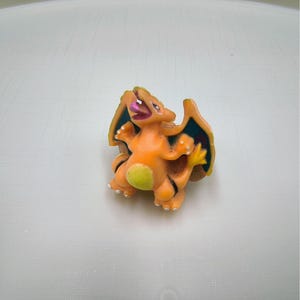 May include: An orange and yellow figurine of a dragon-like creature with a green belly and dark blue wings. The figurine has a red tongue and is posed in a dynamic stance. The toy is on a white surface.