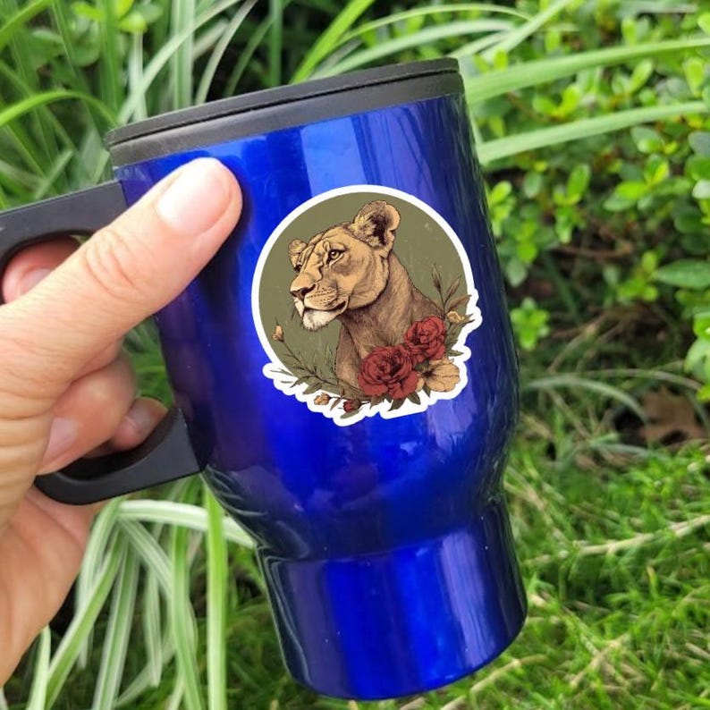 May include: A blue travel mug with a black handle and lid. The mug features a sticker with a lioness illustration, surrounded by red roses and greenery. The lioness is facing left, with a detailed depiction of her face.