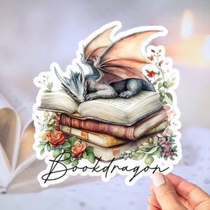 May include: A sticker featuring a watercolor illustration of a blue and gray dragon sleeping on a stack of books. The dragon has a pink and white wing and is surrounded by green leaves and pink flowers. The sticker says "Bookdragon" in a black cursive font.