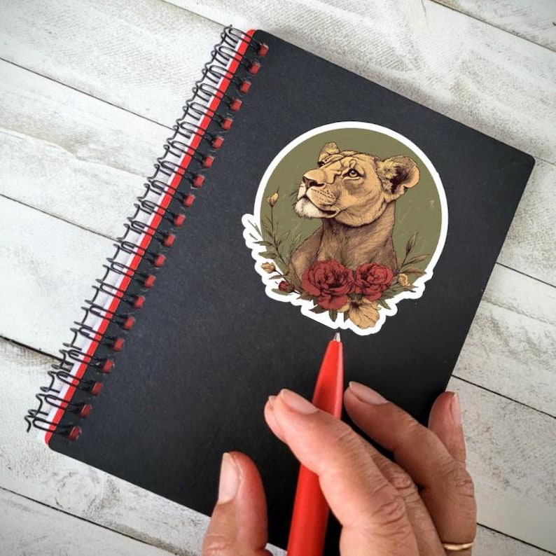 May include: Black spiral-bound notebook with a red spine. A sticker of a lioness with red roses and green foliage is on the cover. A red pen is in the frame. The notebook is on a white wooden surface.