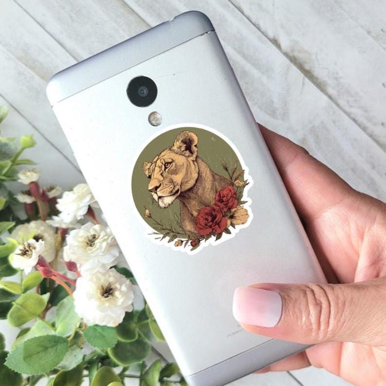 May include: A round sticker featuring a lioness portrait with a floral design. The lioness is depicted in shades of brown and tan, surrounded by red roses and green foliage, set against a green background. The sticker is affixed to a silver smartphone.