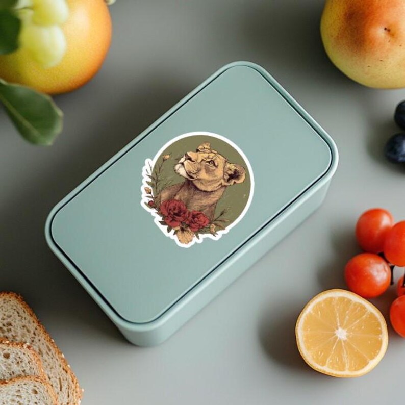 May include: A light teal rectangular lunchbox with a sticker featuring a lioness surrounded by flowers. The lioness is brown and tan, with red and green floral accents. The lunchbox is closed and sits on a gray surface.
