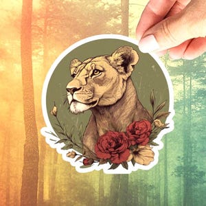 May include: A sticker featuring a detailed illustration of a lioness's head, surrounded by red roses and greenery, set against a green circle. The sticker is held by a hand against a forest backdrop, showcasing a blend of nature and art.