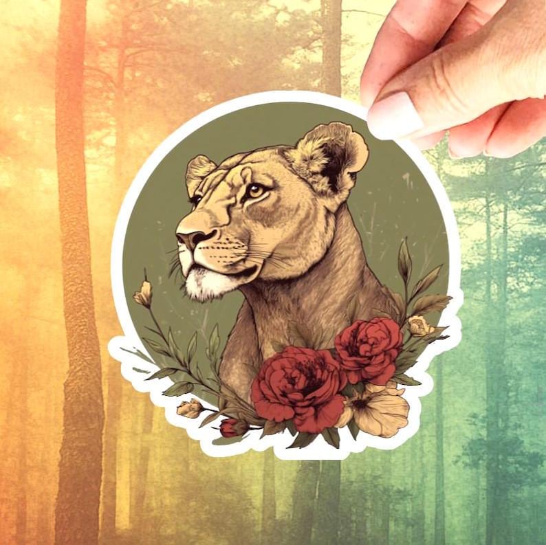 May include: A sticker featuring a detailed illustration of a lioness's head, surrounded by red roses and greenery, set against a green circle. The sticker is held by a hand against a forest backdrop, showcasing a blend of nature and art.