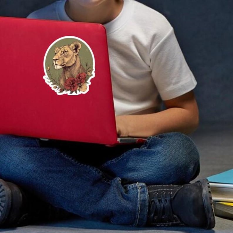 May include: A red laptop with a sticker featuring a lioness head surrounded by flowers. The lioness is depicted in shades of brown and tan, with red and green floral accents. The laptop is on a person's lap.