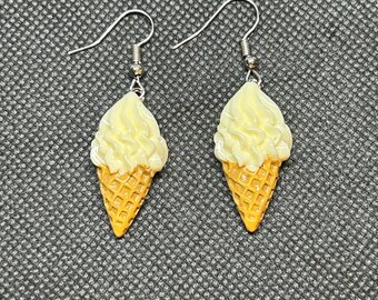 May include: A pair of dangle earrings featuring ice cream cone designs. Each earring has a yellow waffle cone topped with a swirl of creamy, light yellow ice cream. The earrings hang from silver-colored fish hook ear wires.