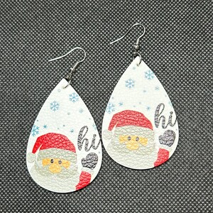 Puede incluir: Par de pendientes en forma de lágrima con un diseño de Papá Noel. Los pendientes tienen un fondo blanco con copos de nieve azules y la palabra "hi". Papá Noel lleva un gorro rojo y tiene barba gris.