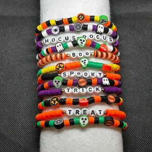 May include: A stack of Halloween-themed bracelets. Each bracelet features a variety of colors, including orange, black, purple, and green. Some bracelets have the words "Hocus Pocus", "Boo", "Spooky", "Trick", and "Treat" spelled out with white letter beads.