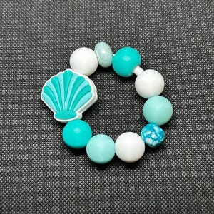 Puede incluir: Una pulsera circular con un dije de concha y una mezcla de cuentas blancas, turquesas y celeste. El dije de concha es blanco con detalles turquesa. Las cuentas son de varios tamaños y texturas, creando un diseño juguetón.