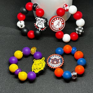 Op de afbeelding: Vier armbanden met kralen en teamthema-bedels. De armbanden hebben rode, zwarte, witte, gele, paarse, oranje en blauwe kralen. Bedels zijn onder meer een bulldog, een logo met de letter "A", een tijger en een logo met de letter "A".