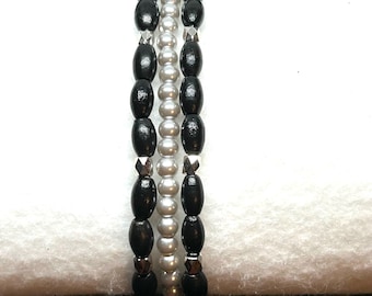 May include: Three beaded bracelets. Two bracelets feature black oval beads, while the centre bracelet has silver-coloured round beads. The bracelets are strung together with a stretchy cord, creating a layered look. The beads are arranged in a repeating pattern.