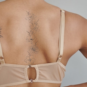 May include: A woman's back with a delicate floral tattoo of a vine with flowers and leaves. The tattoo is on the upper back and extends down to the lower back. The woman is wearing a nude-colored bra with thin straps.