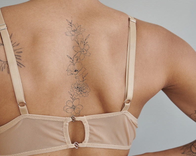 May include: A woman's back with a delicate floral tattoo of a vine with flowers and leaves. The tattoo is on the upper back and extends down to the lower back. The woman is wearing a nude-colored bra with thin straps.