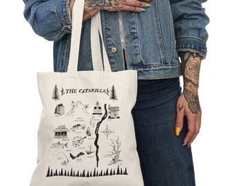 Könnte beinhalten: Eine weiße Canvas-Tragetasche mit schwarzen Illustrationen einer Karte der Catskills, New York. Die Karte enthält Sehenswürdigkeiten, einen Fluss und Bäume. Der Text "THE CATSKILLS" ist oben auf der Tasche in Schwarz gedruckt.