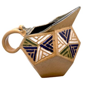 Könnte beinhalten: Eine beige Keramikkrug mit einem geometrischen Design. Der Krug hat einen Griff und einen Ausguss. Das geometrische Design weist blaue, weiße und grüne Akzente auf. Der Krug wird wahrscheinlich zum Servieren von Getränken verwendet.