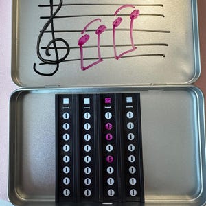May include: An open, rectangular, silver-colored tin with a treble clef and three pink musical notes drawn on the lid. Inside are four black strips with white and pink markings, likely for music notation or education.