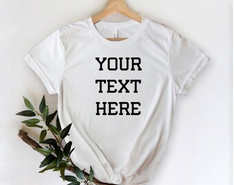 May include: A white t-shirt with the words "YOUR TEXT HERE" printed in a bold, black font. The t-shirt is displayed on a wooden hanger against a white backdrop. A sprig of green leaves and a brown branch are placed near the shirt.