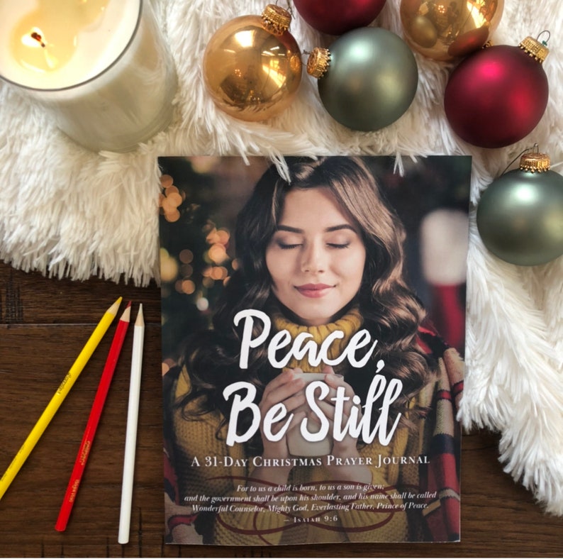 Op de afbeelding: Een 31-daags kerstgebedenboek met een witte achtergrond en een vrouw met haar ogen dicht op de omslag. De tekst op de omslag luidt "Peace, Be Still".