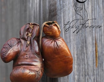 May include: A pair of brown leather boxing gloves hanging on a wooden wall. The gloves are laced up and have a worn, vintage look.
