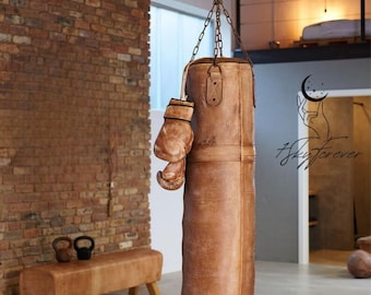 May include: A brown leather boxing bag hanging from a chain with a brown leather boxing glove hanging from the bag. The bag is in front of a brick wall and a wooden bench.