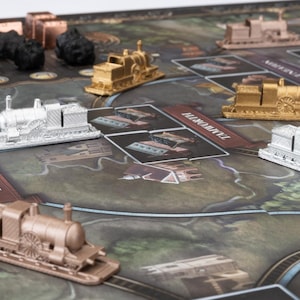 May include: A board game with a map design, featuring miniature train engine game pieces in silver, gold, and bronze colors. The game board includes cards and tokens. The text "BURTON-ON-TRENT" is visible on a card.