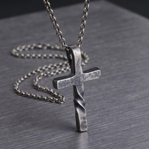 May include: A silver-toned cross pendant necklace. The cross has a twisted lower portion and a simple, classic design. The chain is a matching silver color, and the necklace is displayed against a textured gray background.