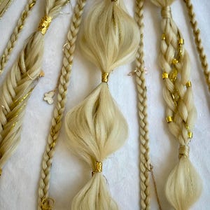 May include: Close-up of blonde hair braids with gold accents. The braids are adorned with gold beads, charms, and decorative bands. The hair is styled in various patterns, including fishtail and classic braids.