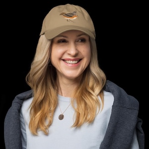 Peut inclure: Une femme portant une casquette de baseball marron clair avec un motif d'oiseau brodé. La casquette a une visière incurvée et une sangle réglable marron. Elle porte un t-shirt bleu clair et une veste gris foncé.