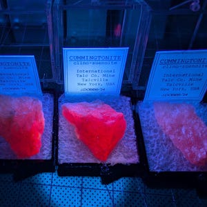 May include: Three specimens of cummingtonite clino-suenoite minerals in clear display cases. The minerals glow bright red under a black light. Each case has a label with the mineral name, origin, and the text "International Talc Co. Mine Talcville New York, USA J.DOBBS '24."