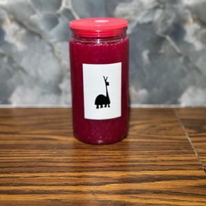 May include: A cylindrical glass jar with a red lid, filled with a deep magenta liquid. A white label with a black llama silhouette is affixed to the jar. The jar sits on a wooden surface.