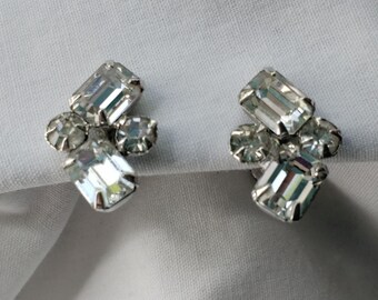 May include: A pair of vintage clip-on earrings featuring clear, rectangular and round-cut rhinestones. The earrings have a silver-tone metal setting and are displayed on a white fabric background. The design is a cluster of sparkling stones.