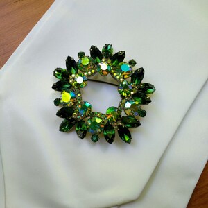 May include: A circular brooch with a wreath design, featuring emerald green and iridescent crystals. The brooch is set on a white fabric, with a wooden surface in the background. The crystals have a variety of shapes and sizes.