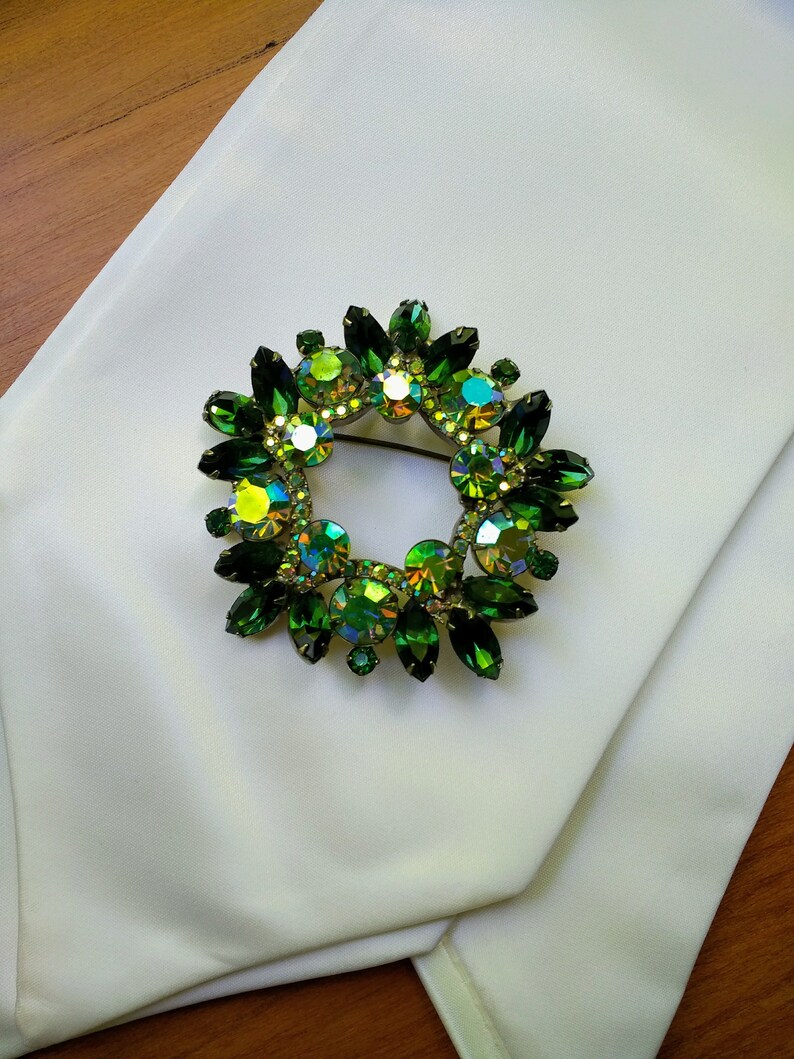 May include: A circular brooch with a wreath design, featuring emerald green and iridescent crystals. The brooch is set on a white fabric, with a wooden surface in the background. The crystals have a variety of shapes and sizes.
