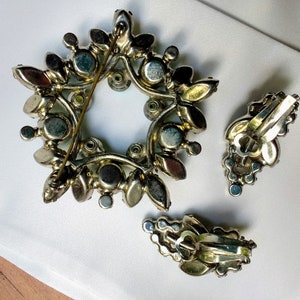 May include: Vintage jewelry set featuring a starburst brooch and matching clip-on earrings. The brooch and earrings are silver-toned metal with a floral design, adorned with iridescent and clear rhinestones. The brooch measures approximately 2 inches in diameter.
