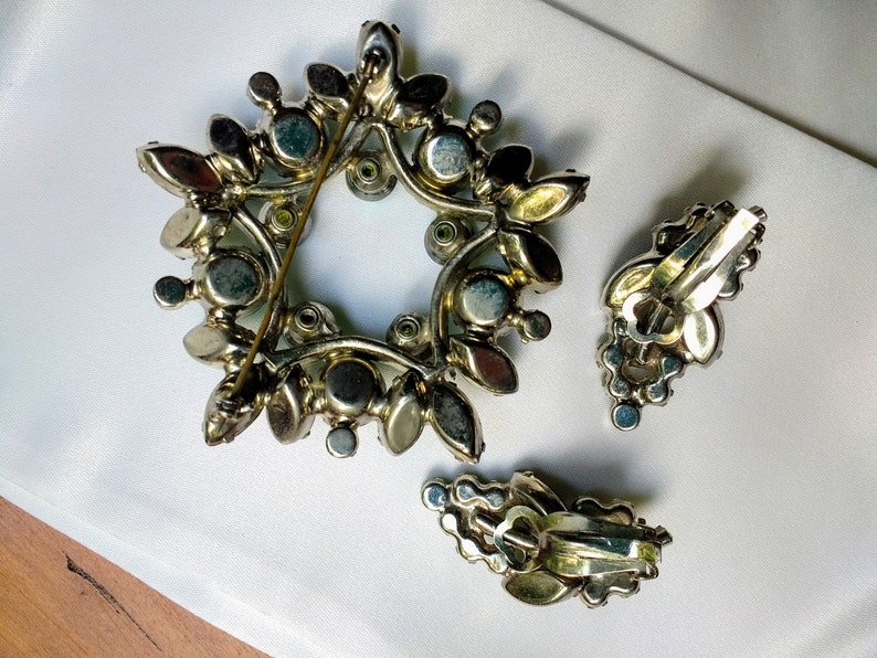 May include: Vintage jewelry set featuring a starburst brooch and matching clip-on earrings. The brooch and earrings are silver-toned metal with a floral design, adorned with iridescent and clear rhinestones. The brooch measures approximately 2 inches in diameter.