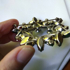 May include: Vintage silver-toned brooch with a floral design. The brooch features a wreath-like arrangement of clear and amber-colored rhinestones. The pin is held in place by a safety clasp.